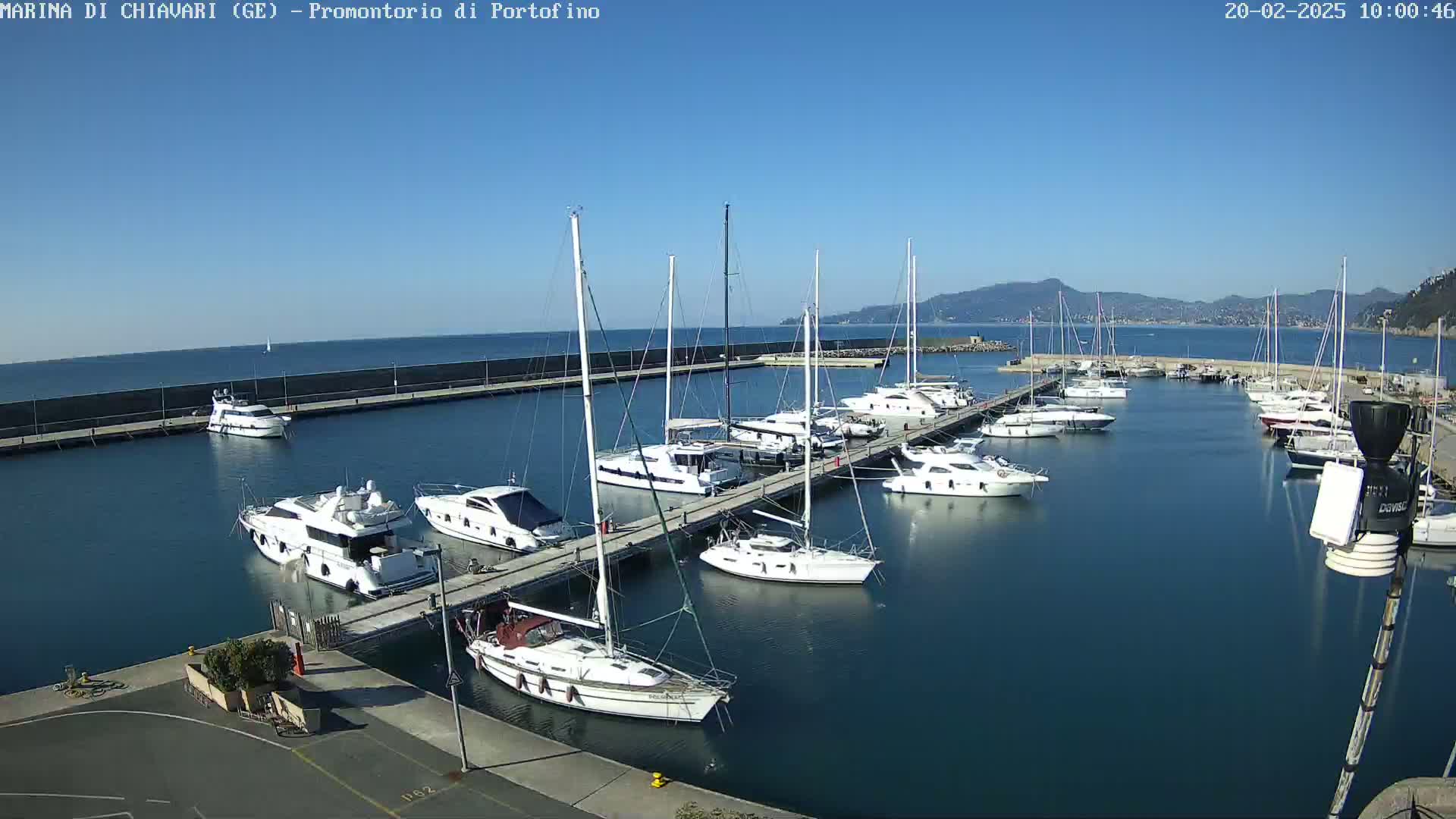 A marina filled with numerous yachts and sailboats on a sunny day, with calm blue water and distant mountains visible in the background.
