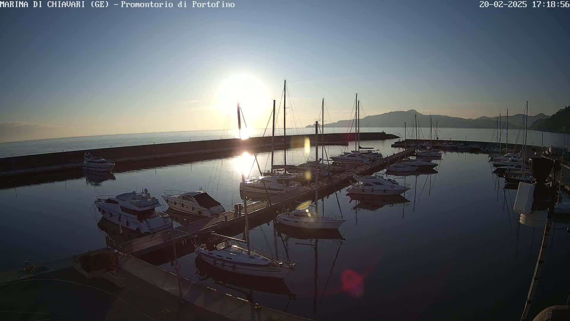 Numerous sailboats and motorboats are docked in a marina at sunset, with calm water reflecting the boats and the setting sun.