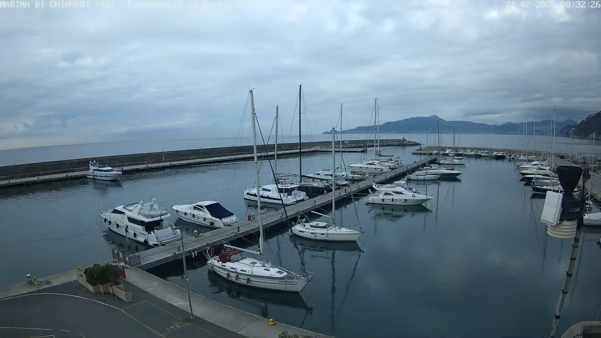 A marina filled with yachts and sailboats on a cloudy day, nestled between a sea wall and a distant mountain range.