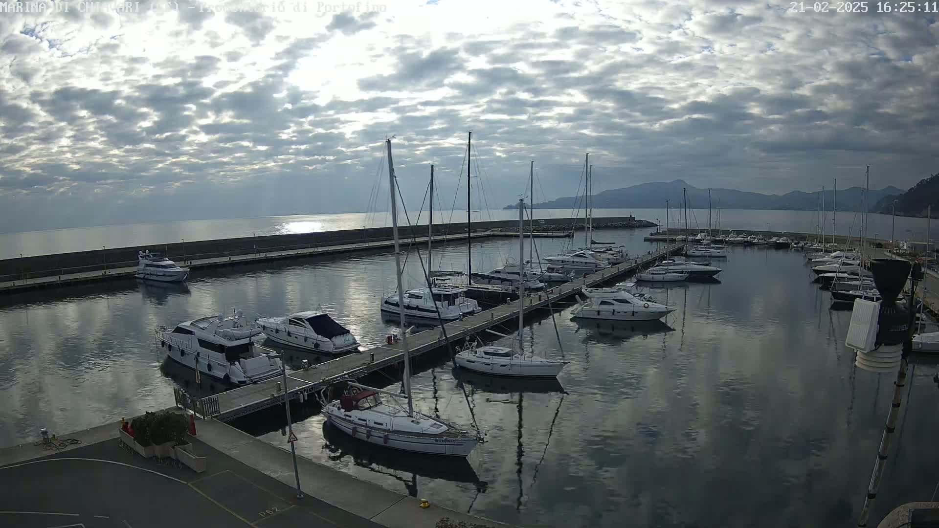 A marina filled with sailboats and motorboats is seen under a partly cloudy sky, with calm water reflecting the clouds and boats.