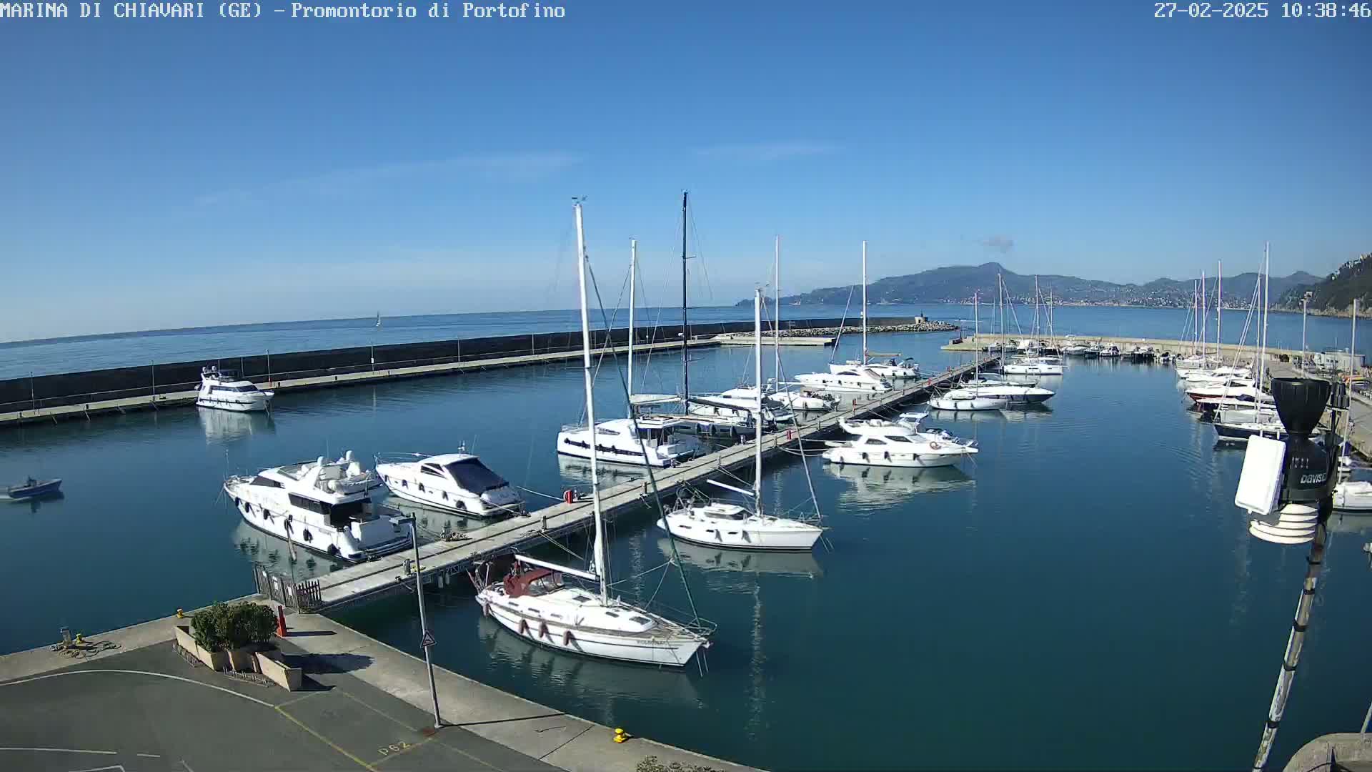 A marina filled with sailboats and motorboats on a sunny day with calm waters and mountains visible in the distance.