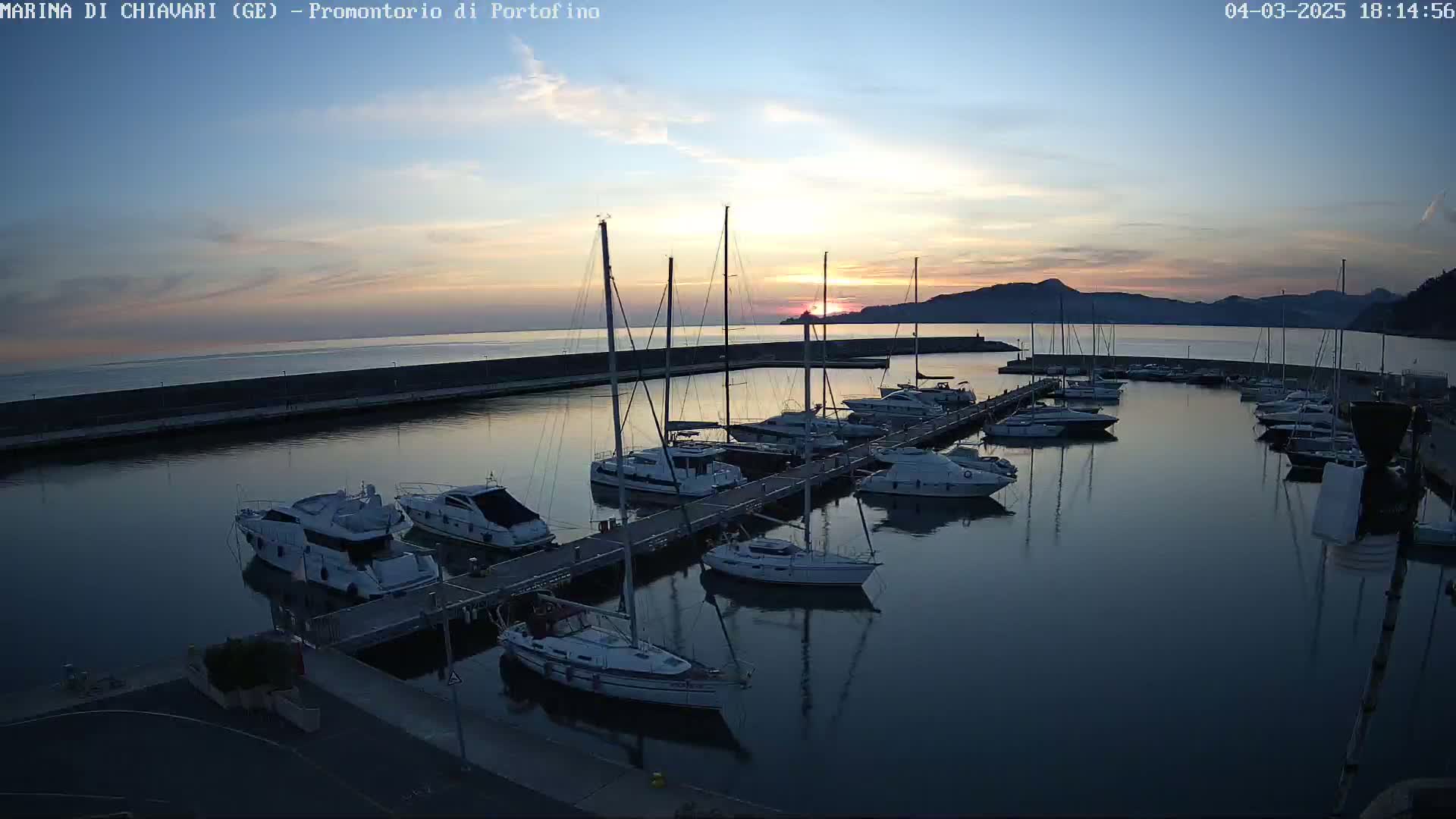 A calm harbor filled with sailboats and motorboats at sunset, under a mostly clear sky.