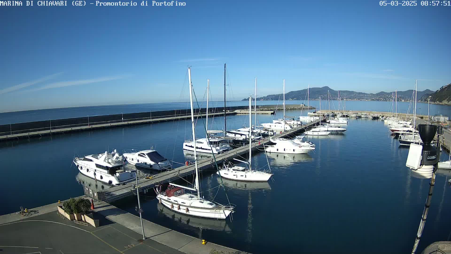 A marina filled with sailboats and motor yachts is situated on calm, clear water under a bright, sunny sky.