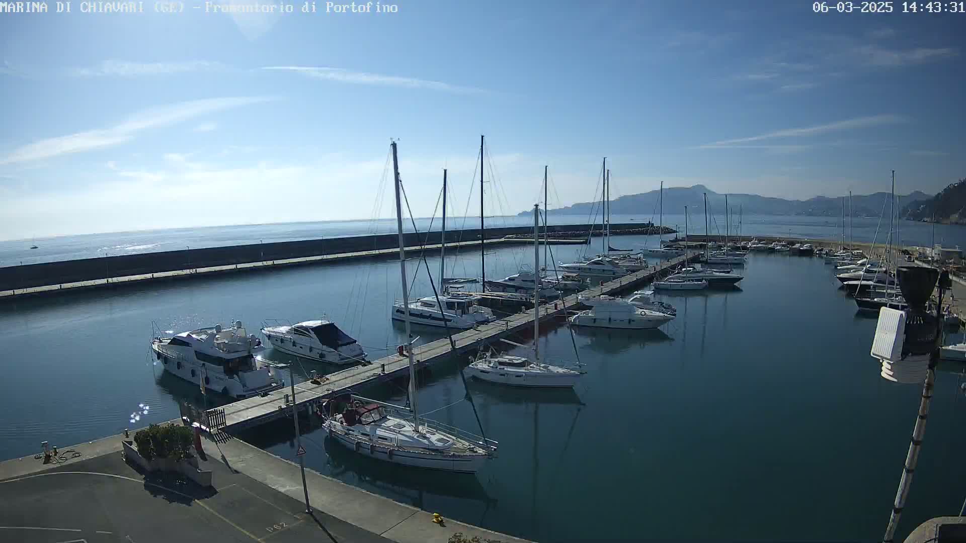 A marina filled with sailboats and motorboats on a sunny day, with calm waters and distant hills visible in the background.