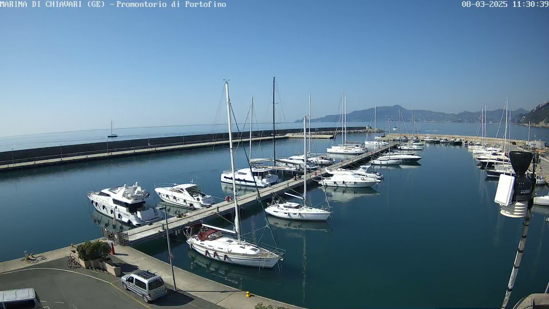 A marina filled with sailboats and motor yachts on a sunny day, with calm water and distant mountains visible in the background.