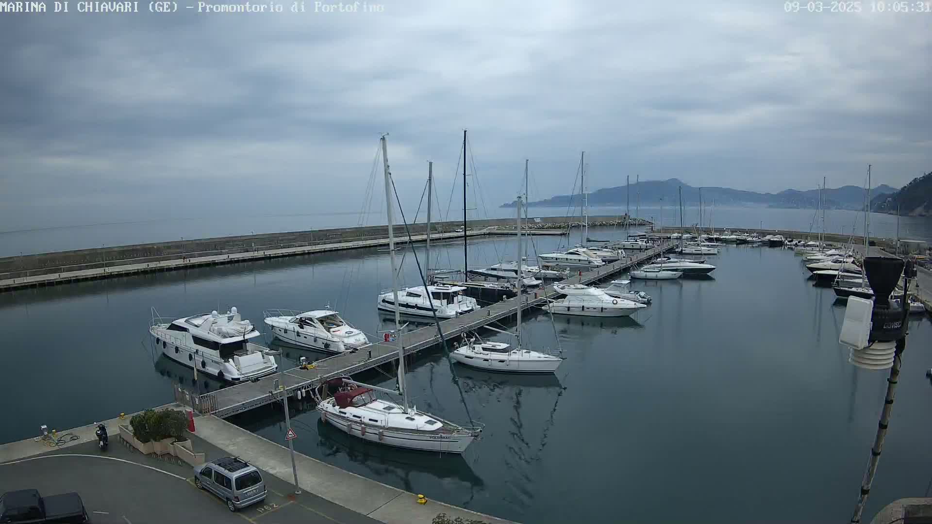 A marina filled with sailboats and motorboats is situated on calm water under a cloudy sky, with mountains visible in the distance.