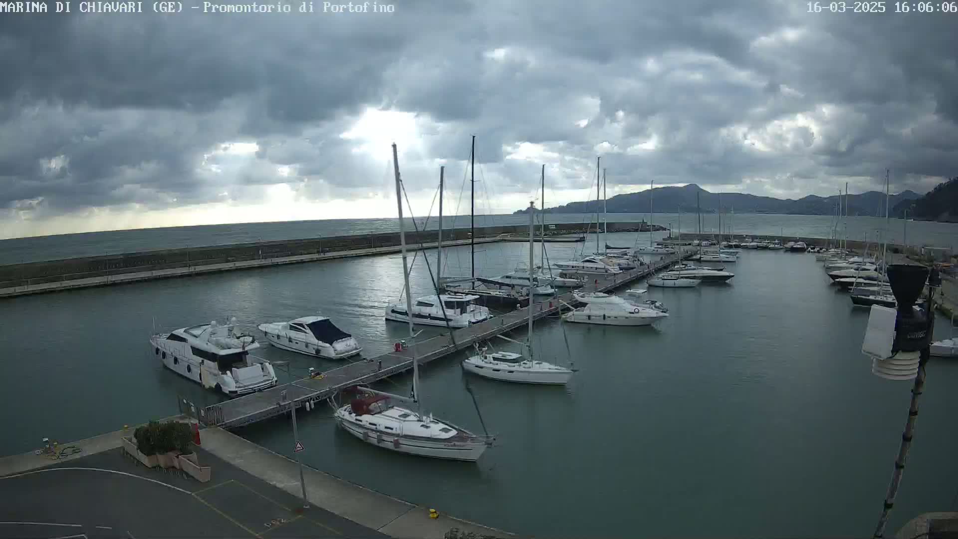 A marina filled with numerous sailboats and motorboats is situated on calm water under a cloudy sky.