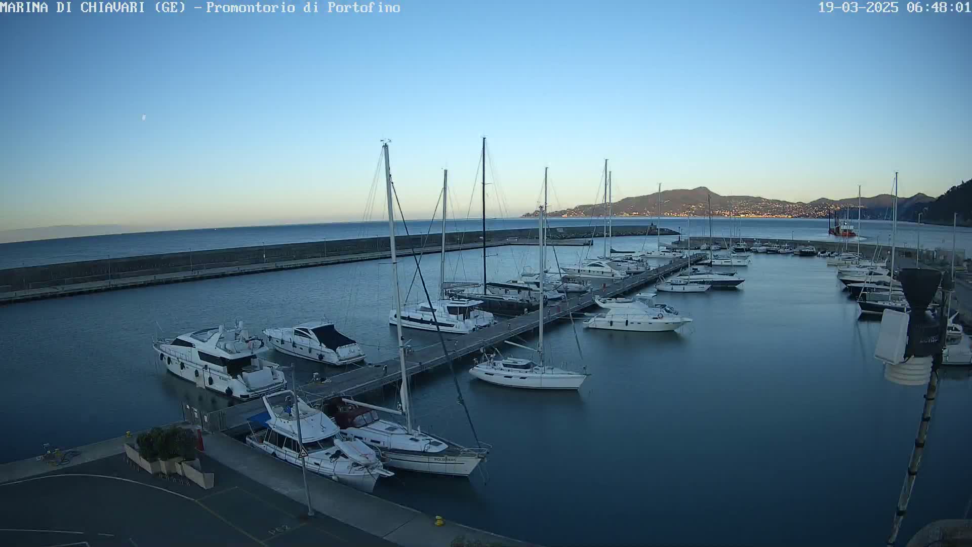 A marina filled with numerous boats and yachts is situated on calm water under a clear, bright blue sky, with a distant mountain range visible in the background.