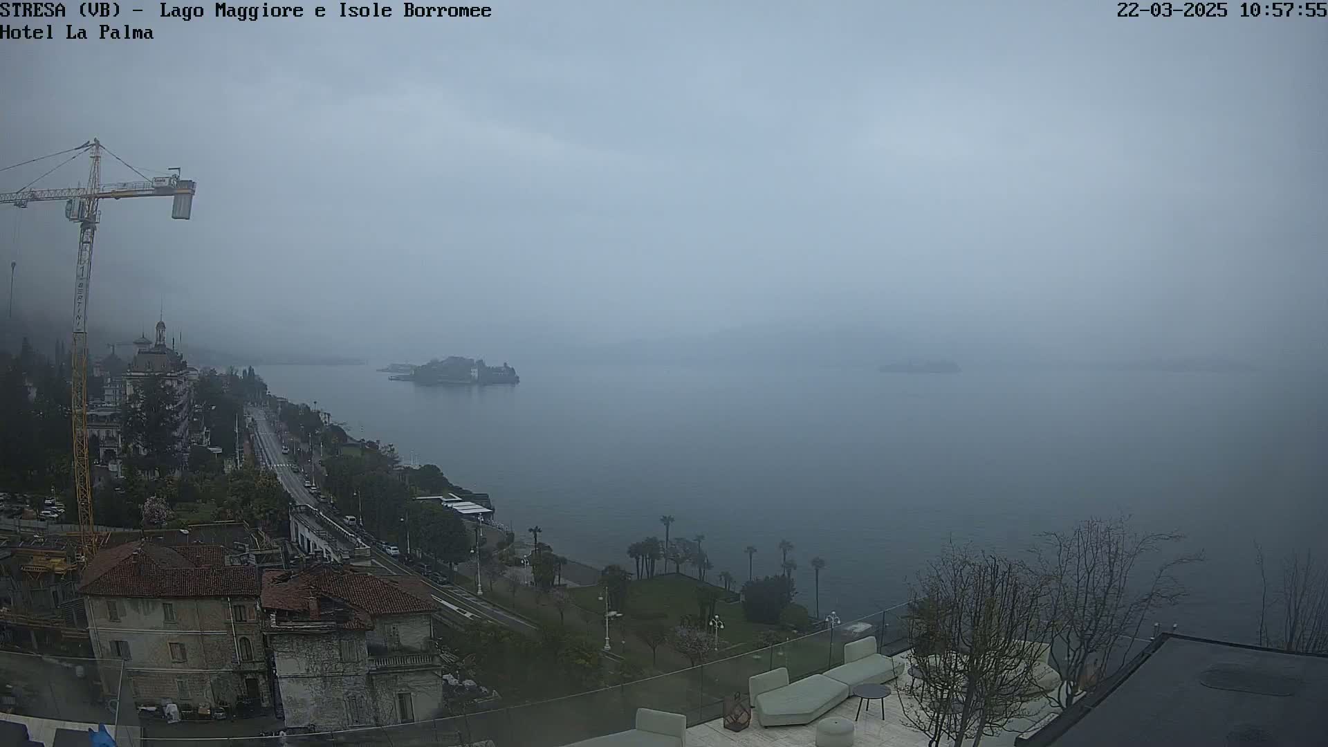 A hazy, overcast view of a lake and shoreline, with buildings, trees, a road, and a construction crane visible.