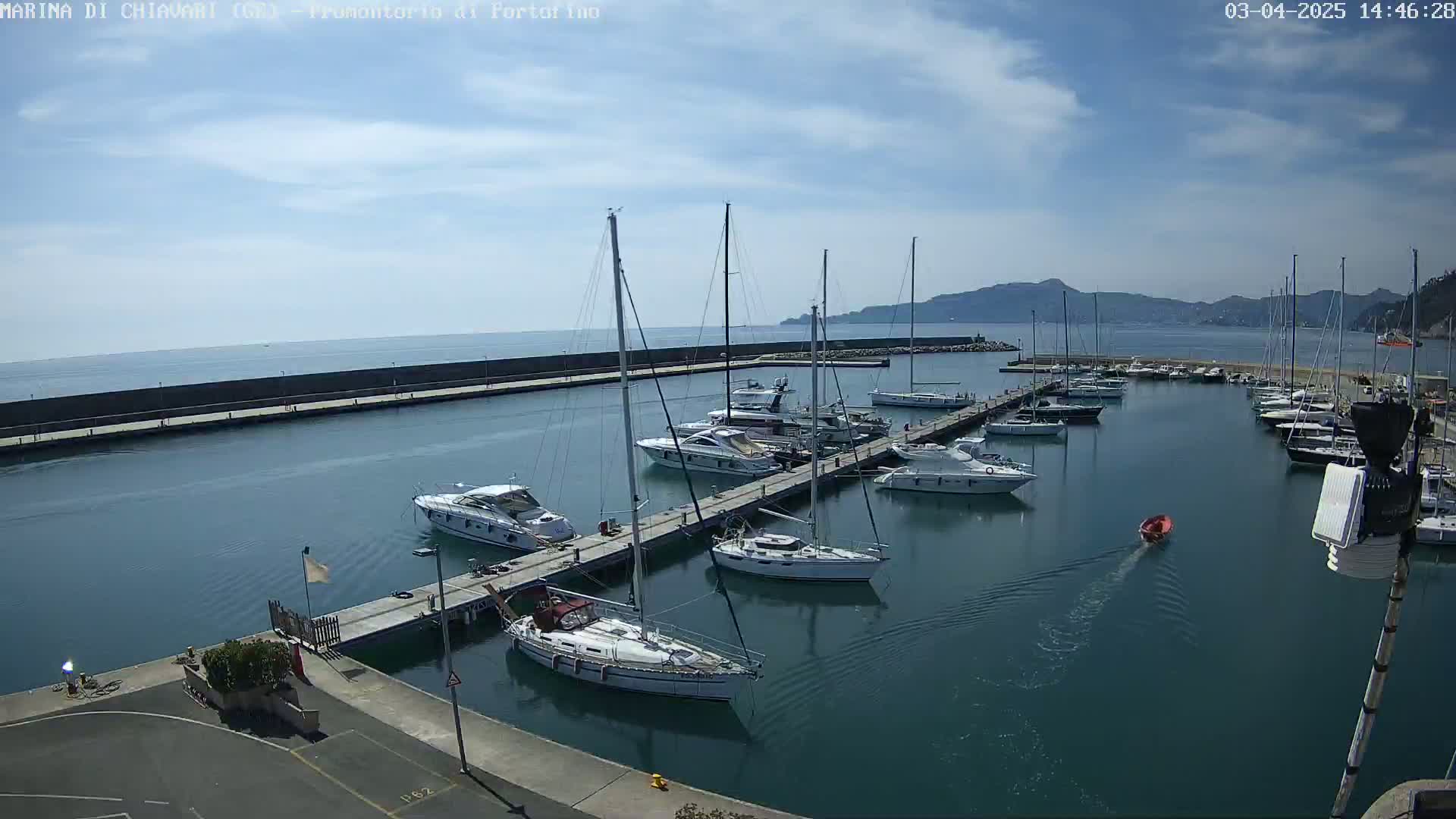 A marina filled with sailboats and motorboats on a partly sunny day, with calm waters and a distant mountain range visible.
