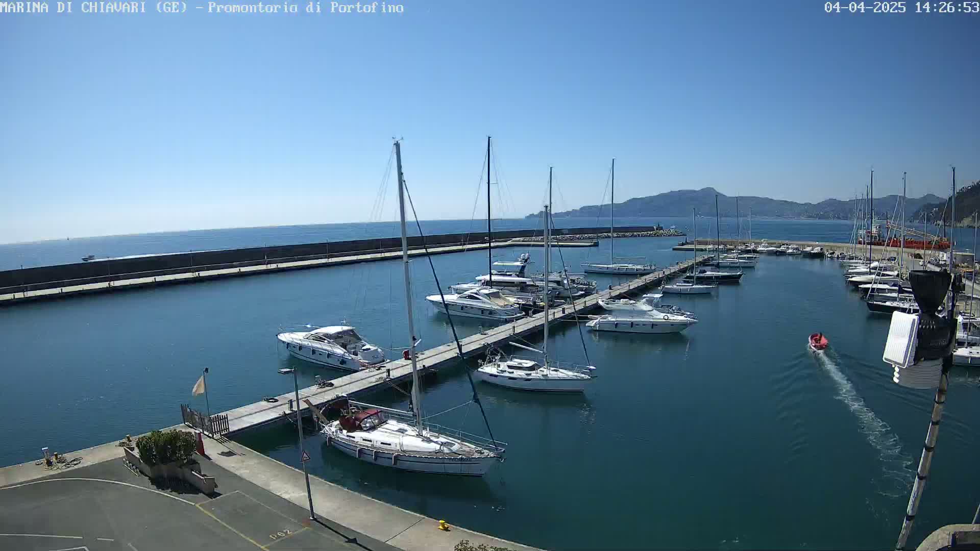 A marina filled with sailboats and motorboats on a sunny day, with calm water and distant hills visible in the background.