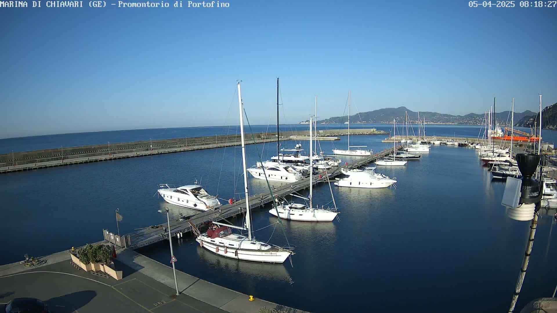 Under a clear blue sky, numerous yachts and sailboats are moored in a marina, with a calm sea stretching to distant hills.