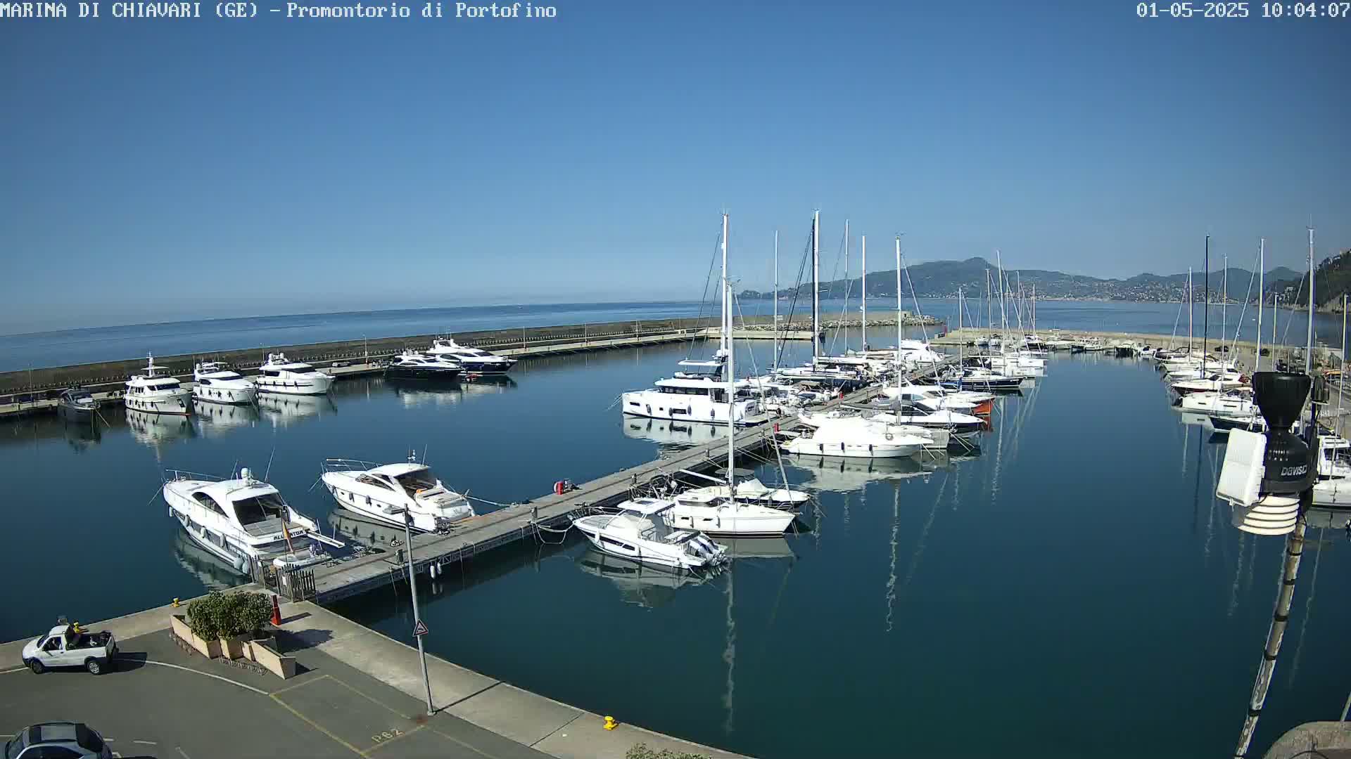 A marina filled with numerous yachts and sailboats on a calm, sunny day, with mountains visible in the distance.