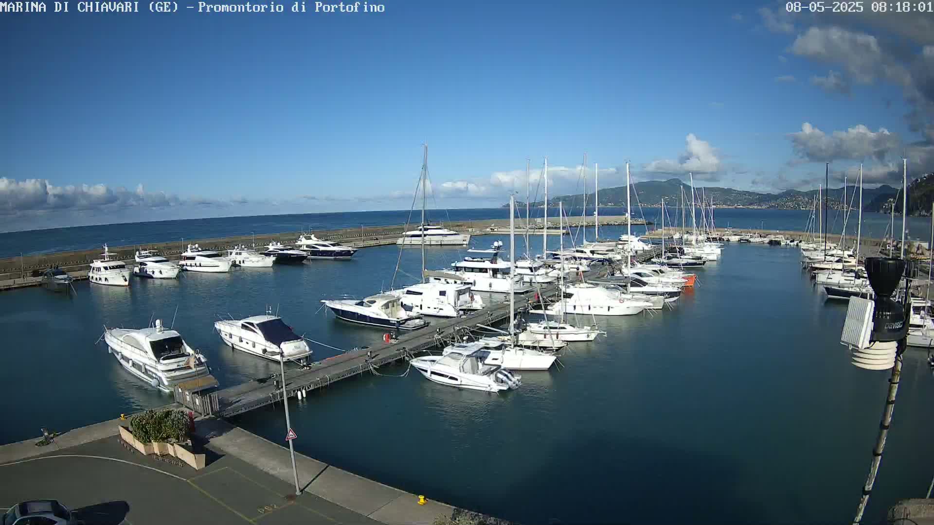 A marina filled with numerous yachts and sailboats is situated on a calm, clear day with a blue sky and a few clouds.