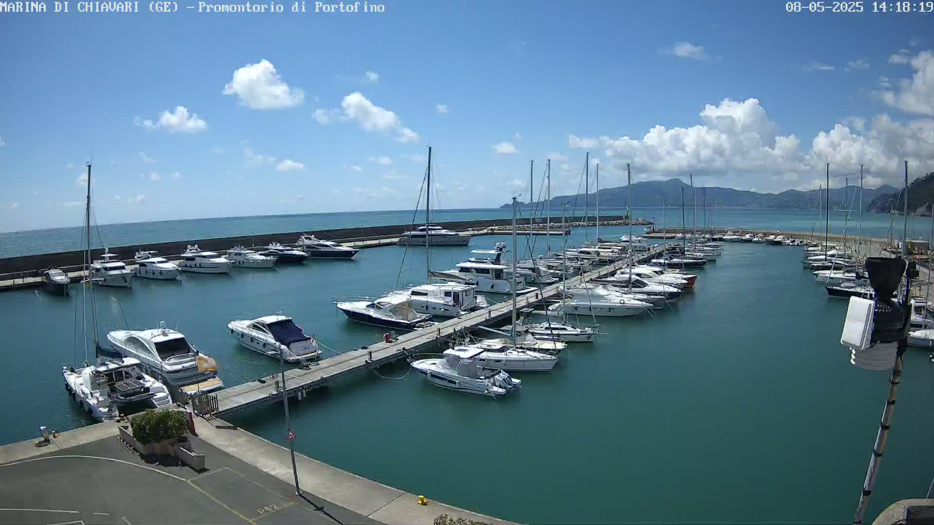A marina filled with numerous yachts and sailboats is situated on a calm, teal body of water under a partly cloudy sky.