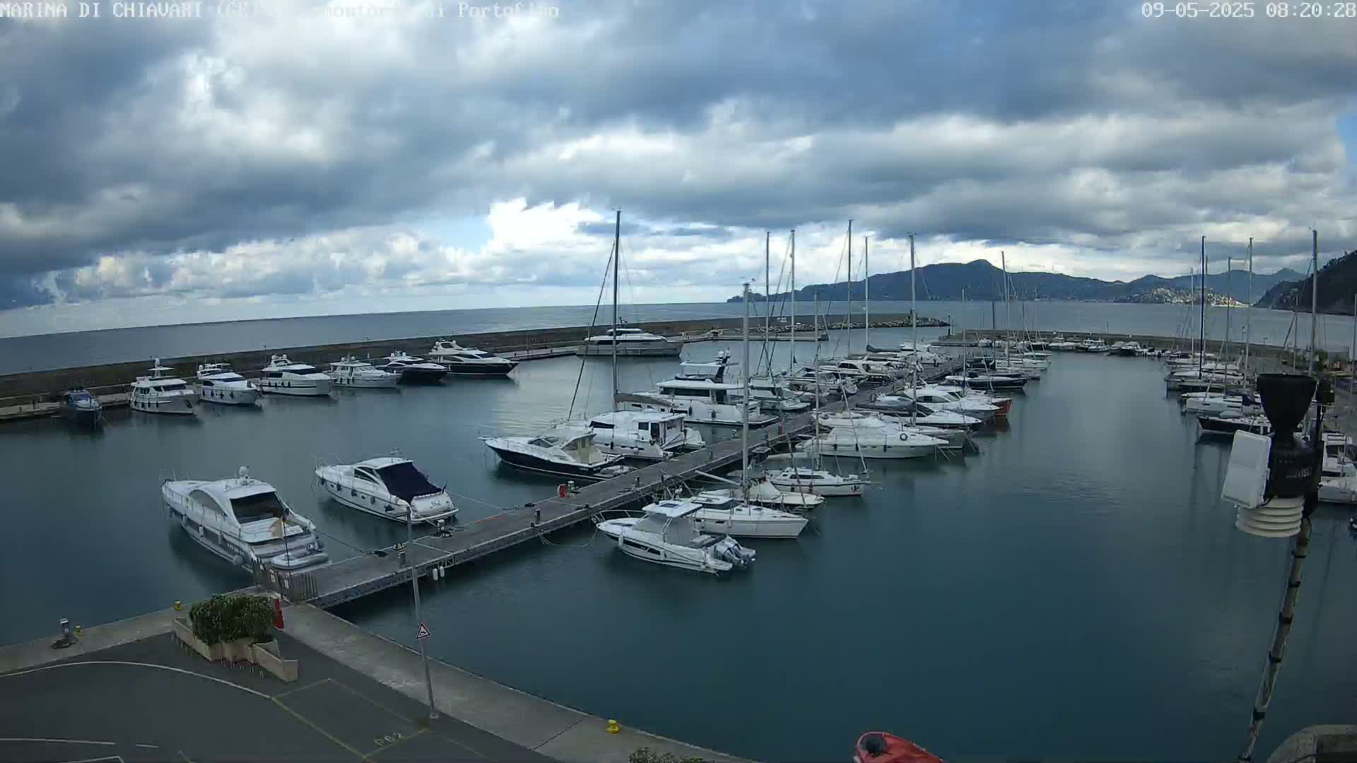 A marina filled with numerous yachts and sailboats is situated on calm, dark-blue water under a cloudy sky.