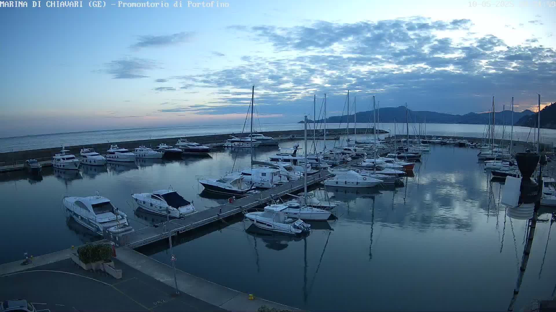 Many boats are docked in a calm marina at dusk under a partly cloudy sky.