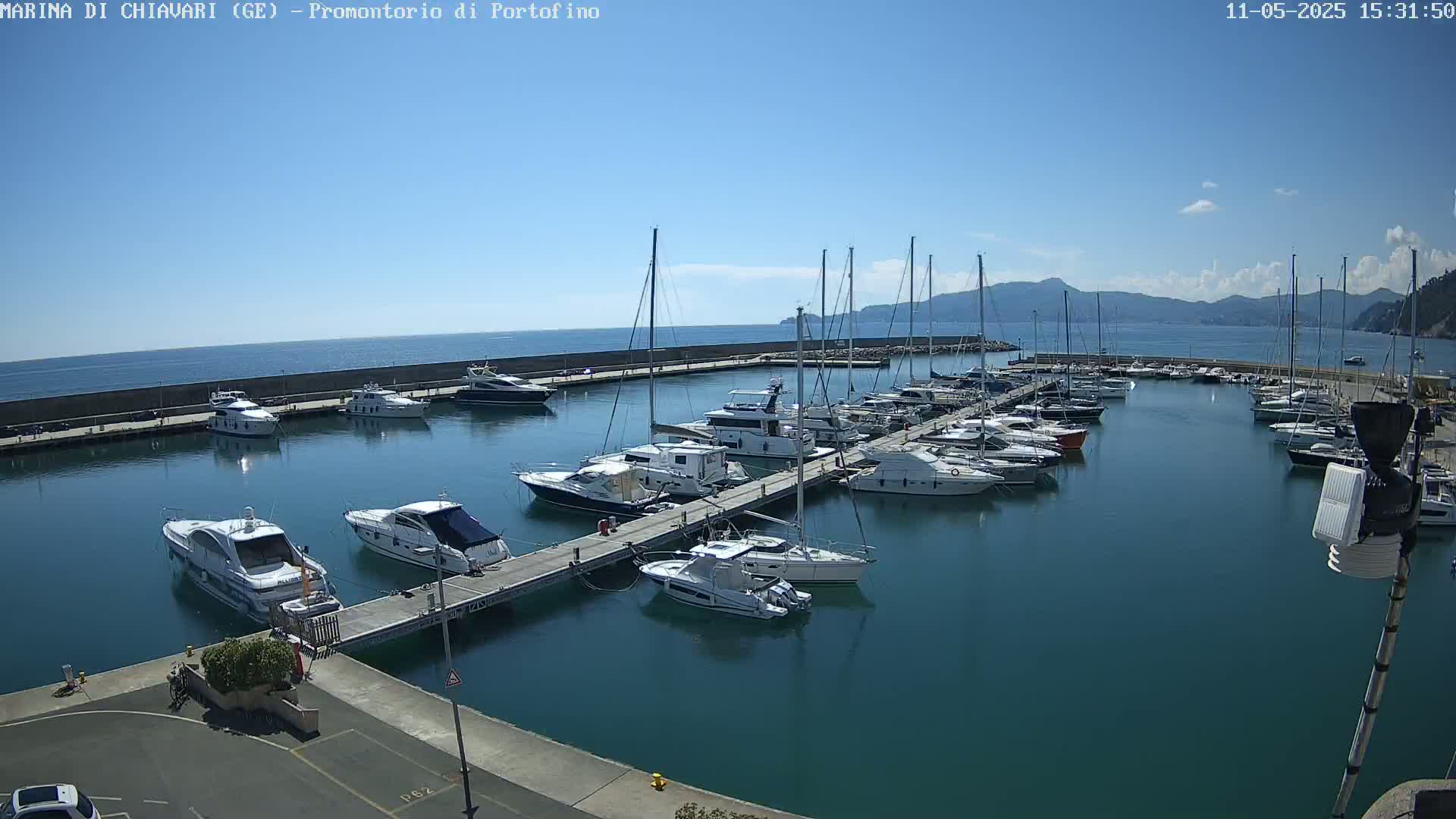 A marina filled with numerous yachts and sailboats is situated on a calm, sunny day with clear blue skies and distant mountains visible in the background.