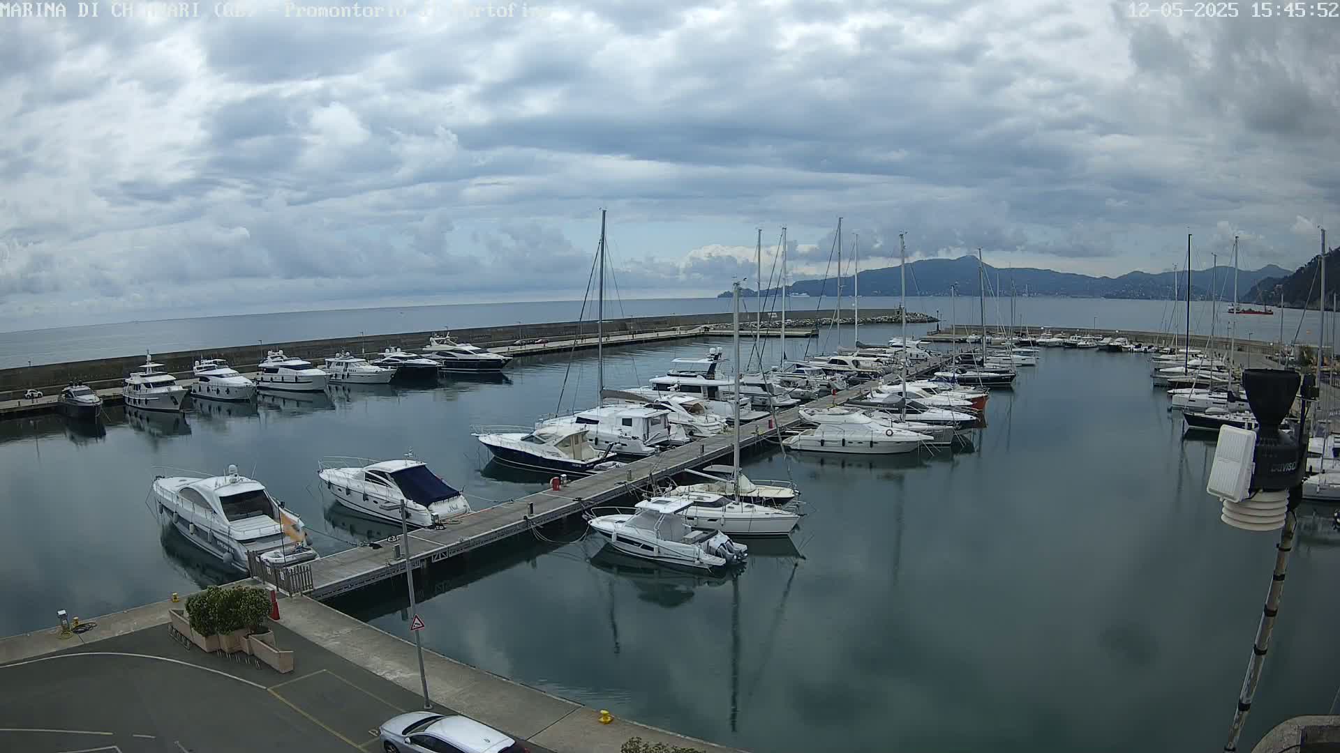 A marina filled with numerous yachts and sailboats is seen on a partly cloudy day, with mountains visible in the distance.