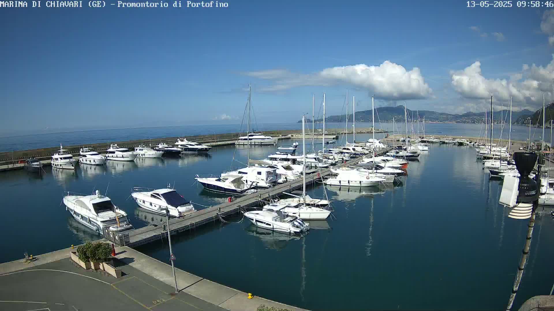 A marina filled with numerous yachts and sailboats is shown on a partly sunny day with a few clouds in the sky.