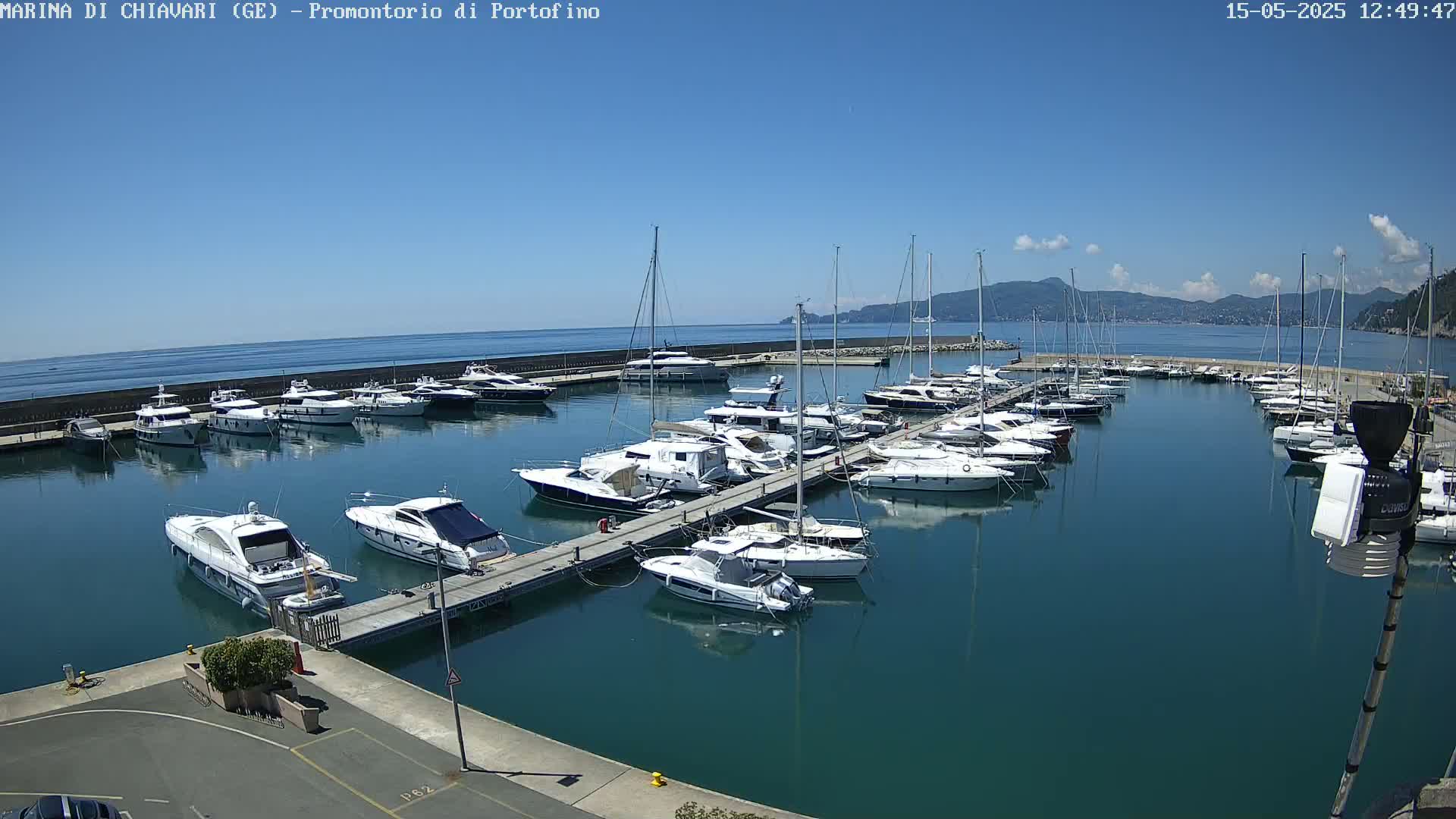 A marina filled with numerous yachts and sailboats is situated on calm, dark-blue water under a mostly clear, sunny sky.