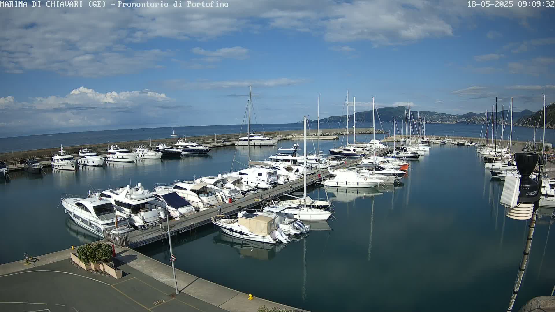 A marina filled with numerous yachts and sailboats is nestled along a calm body of water under a partly cloudy sky.