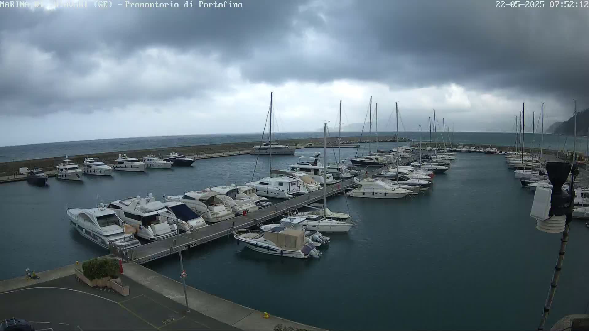 A marina filled with numerous yachts and sailboats is shown under a cloudy, overcast sky with a choppy sea.