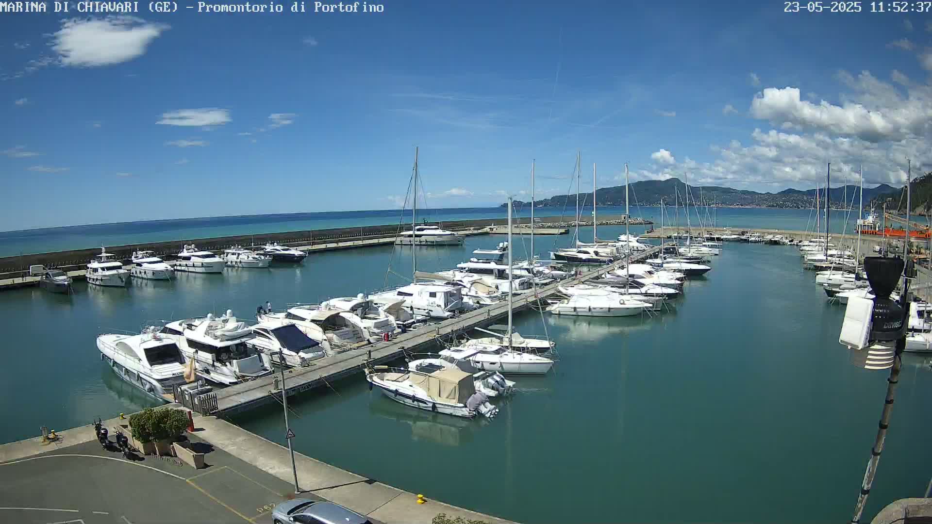 A marina filled with numerous yachts and sailboats is situated on calm, teal water under a mostly sunny sky with some scattered clouds.