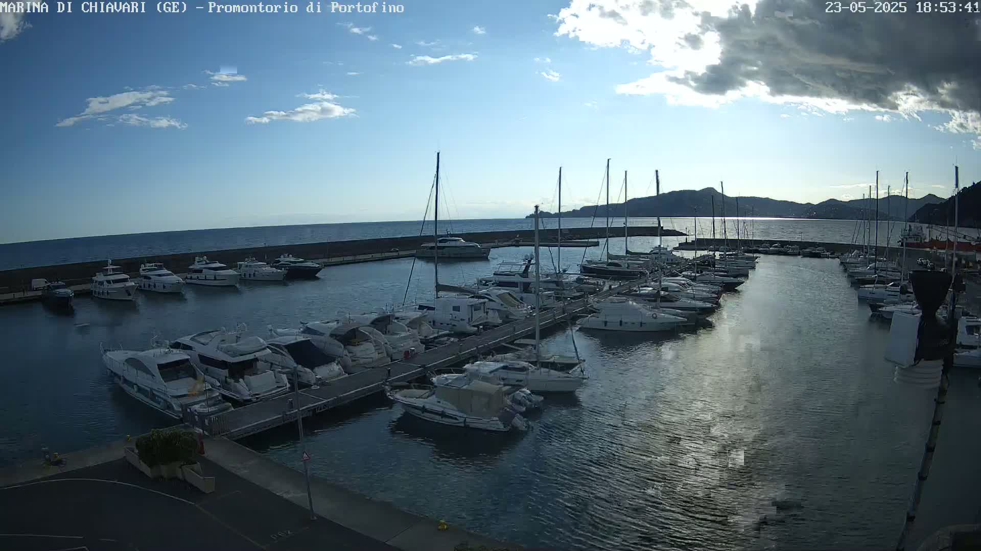 A marina filled with numerous yachts and sailboats is seen under a partly cloudy sky, with calm water reflecting the light.
