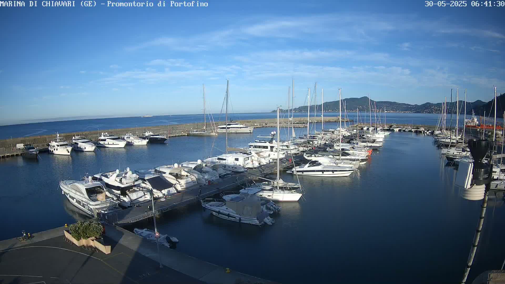 A marina filled with numerous yachts and sailboats is situated on calm, dark blue water under a mostly clear, bright blue sky.