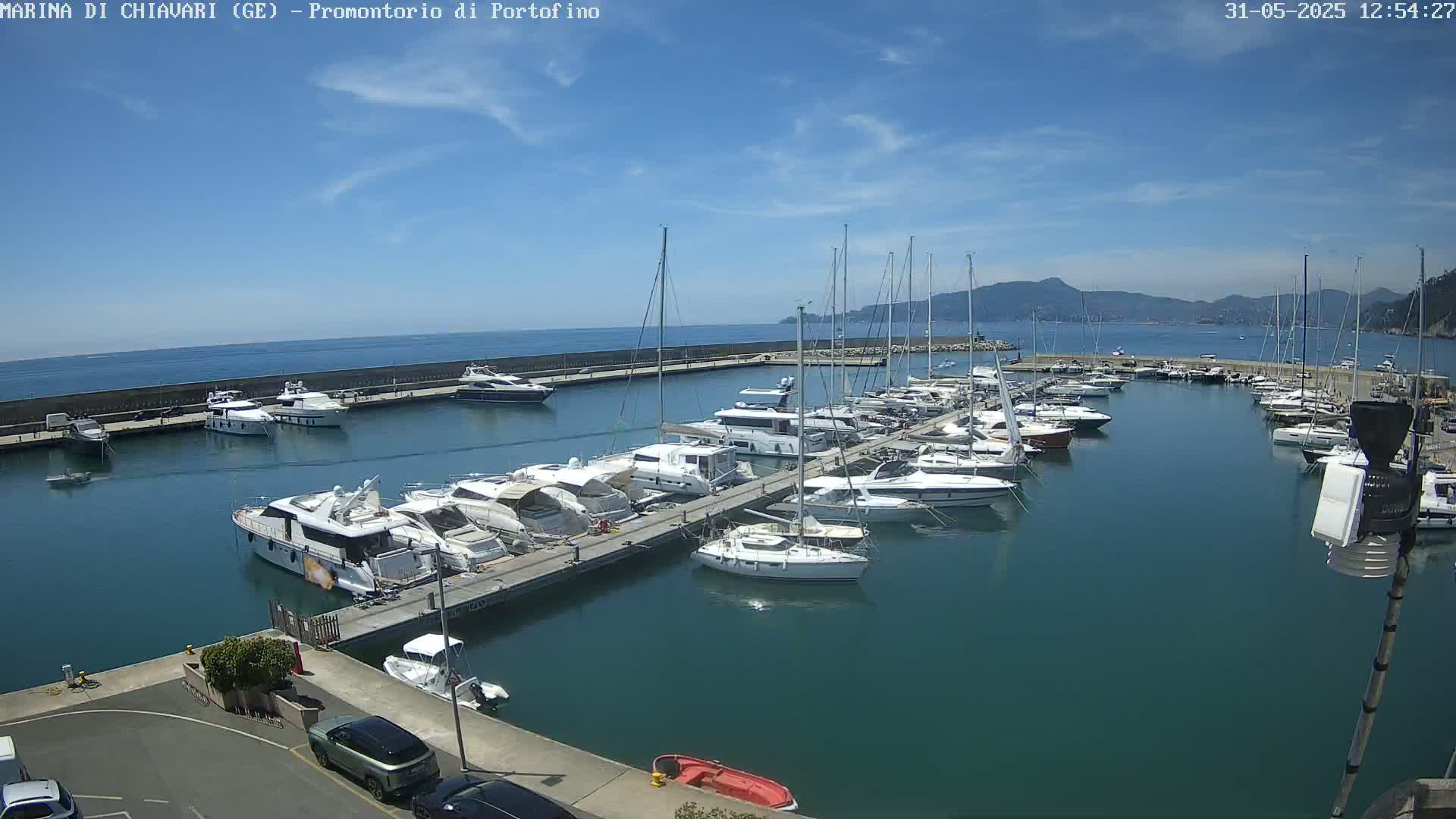 A marina filled with numerous yachts and sailboats is shown on a sunny day with clear blue skies and calm, teal water.