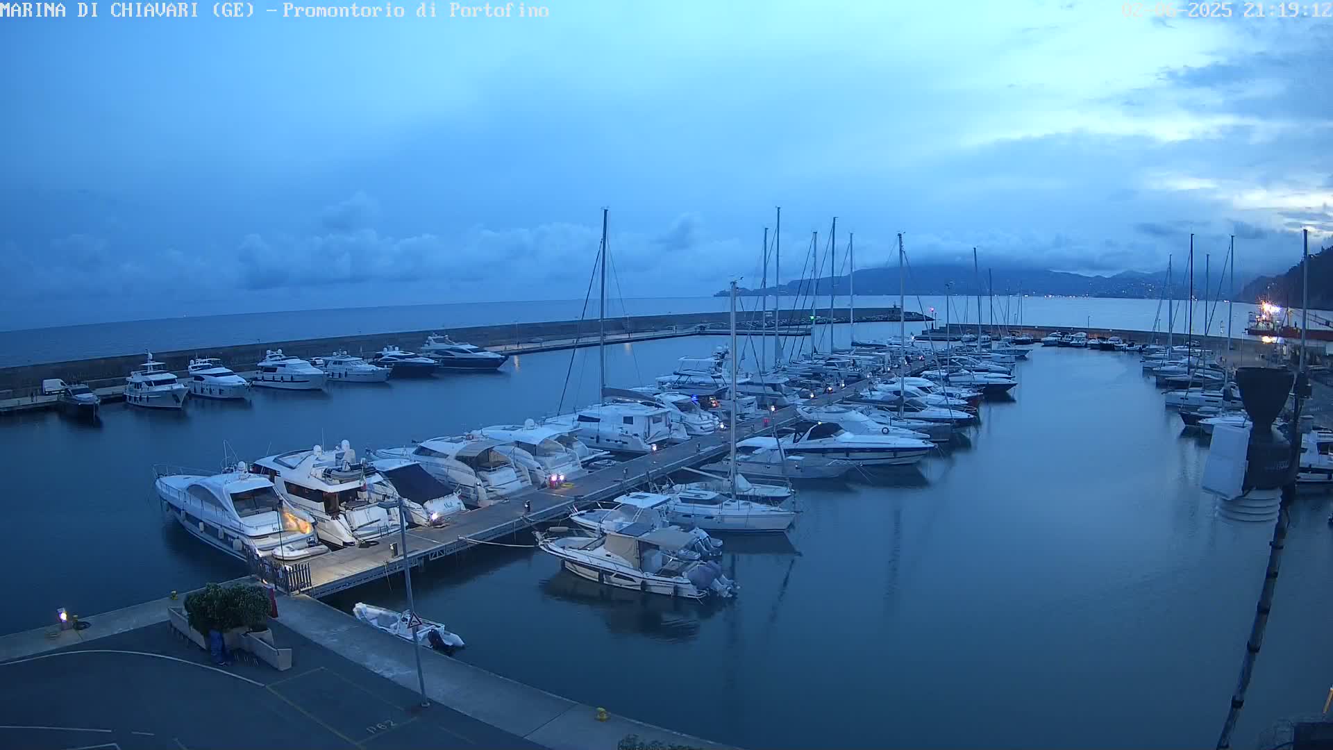 Many yachts and sailboats are moored in a calm harbor at dusk under a mostly cloudy sky.