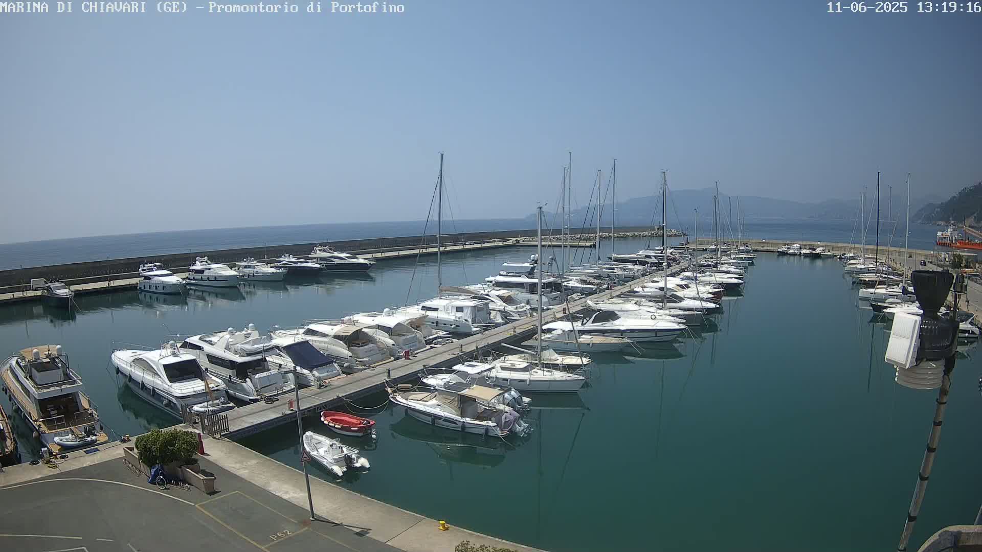 A marina filled with numerous yachts and sailboats is situated on calm, clear water under a bright, sunny sky.