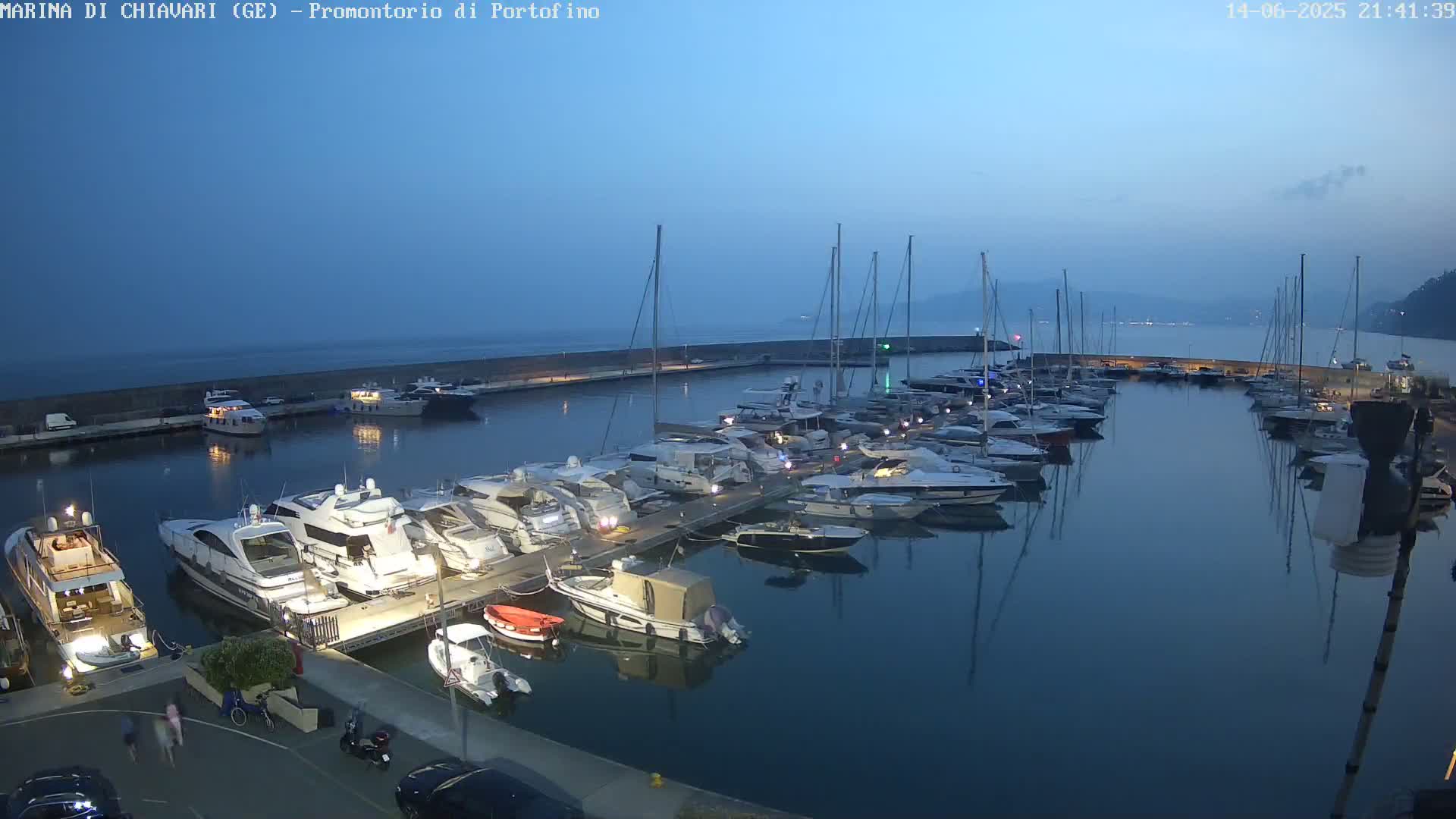 A marina filled with numerous yachts and sailboats is situated on a calm, clear day next to a seawall, with a clear blue sky and distant hills visible in the background.