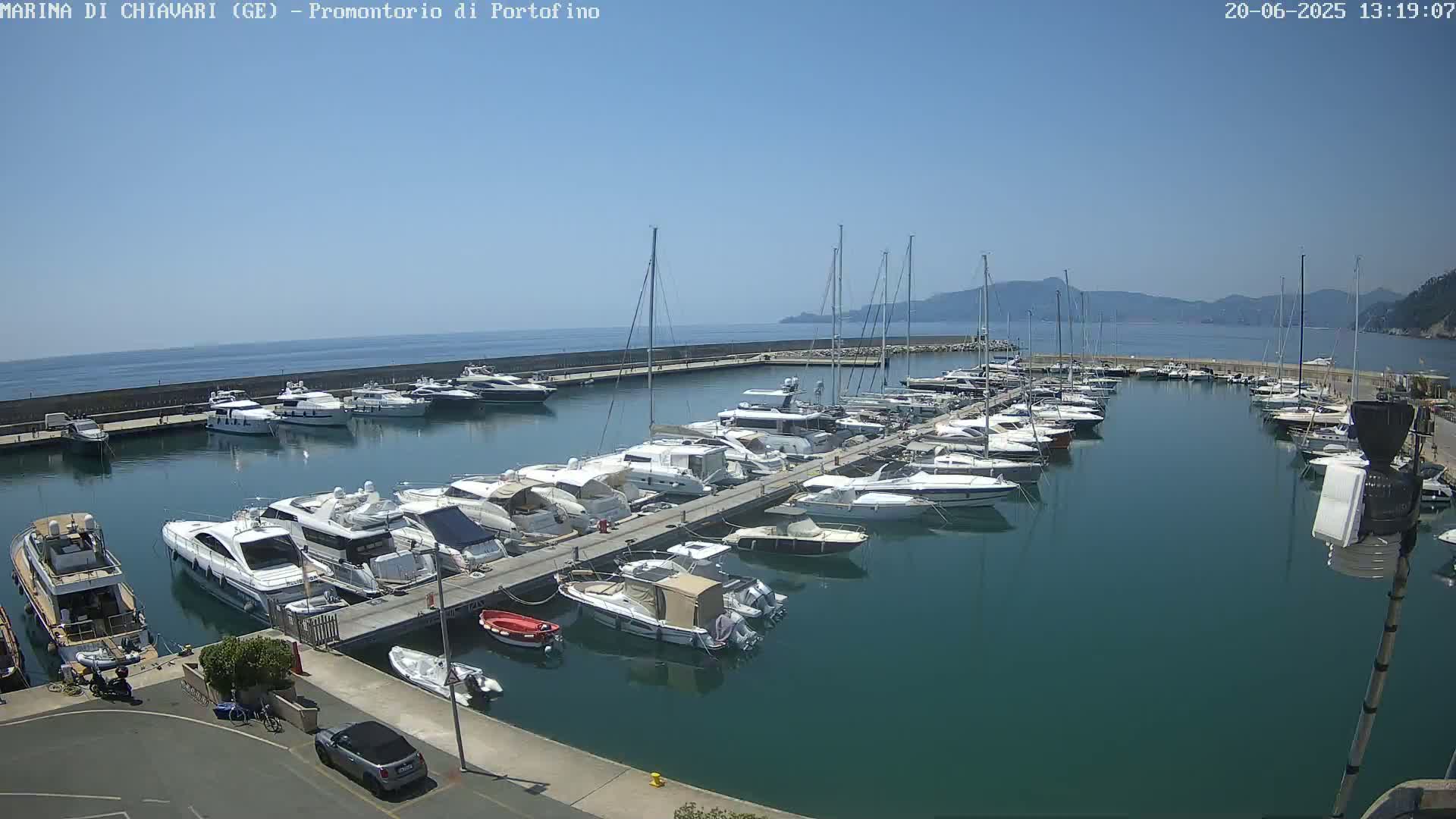 A marina filled with numerous yachts and boats is seen on a sunny day with calm, clear water and distant mountains.