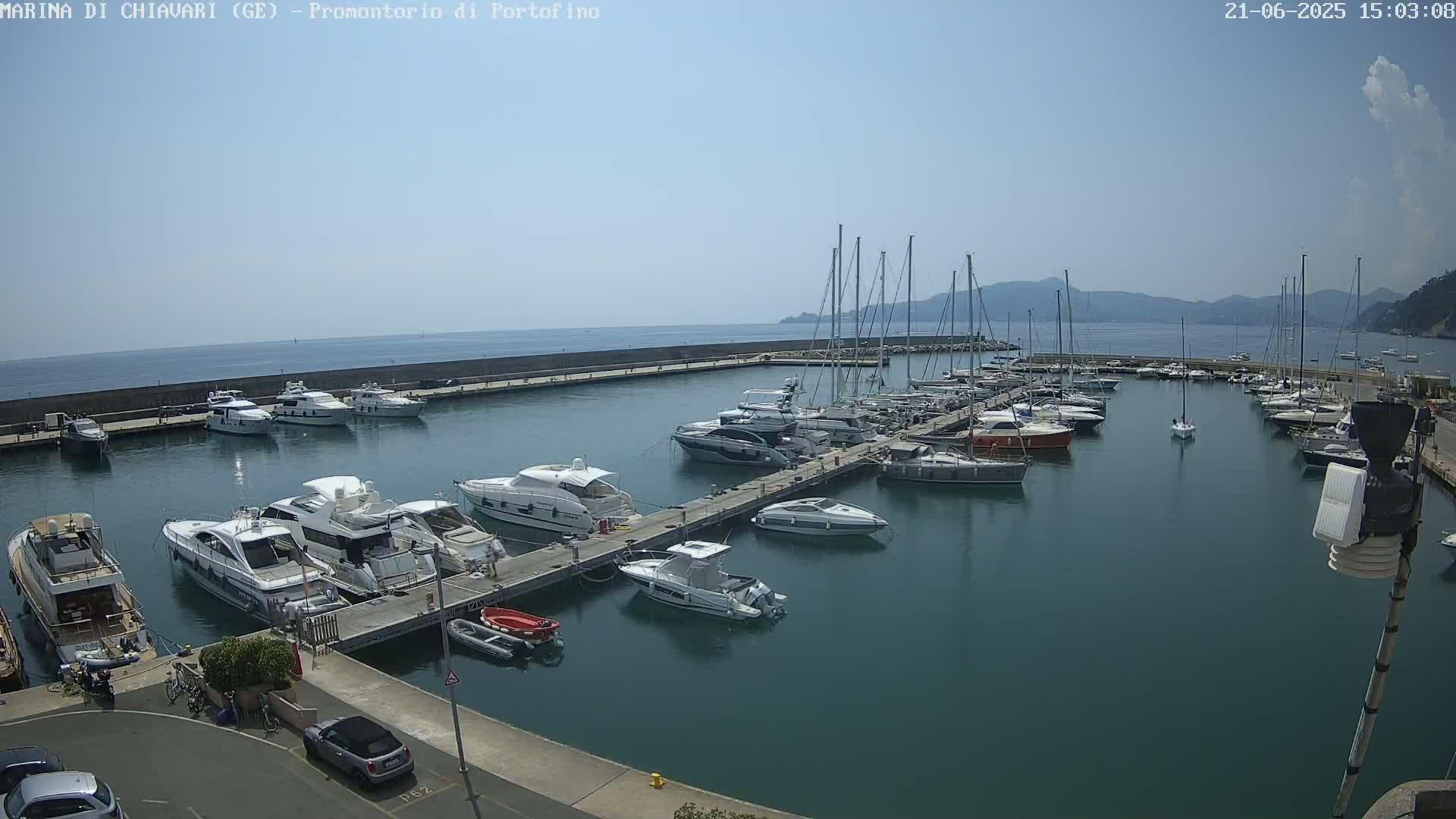 A marina filled with numerous yachts and sailboats is situated on a calm, clear day next to a sea wall overlooking a body of water with distant hills.