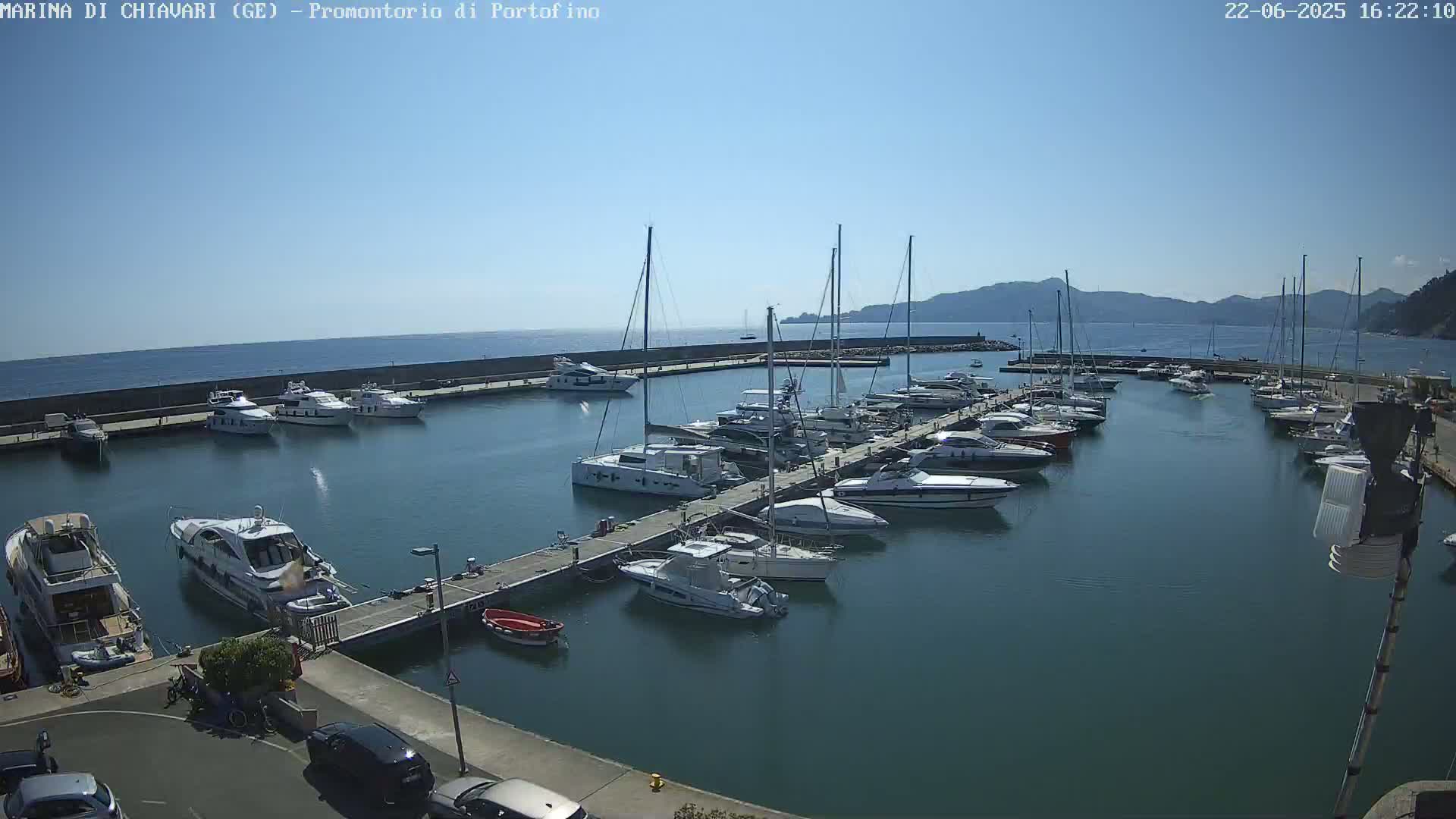 A marina filled with numerous boats and yachts is situated on calm, clear water under a bright, sunny sky, with distant hills visible in the background.