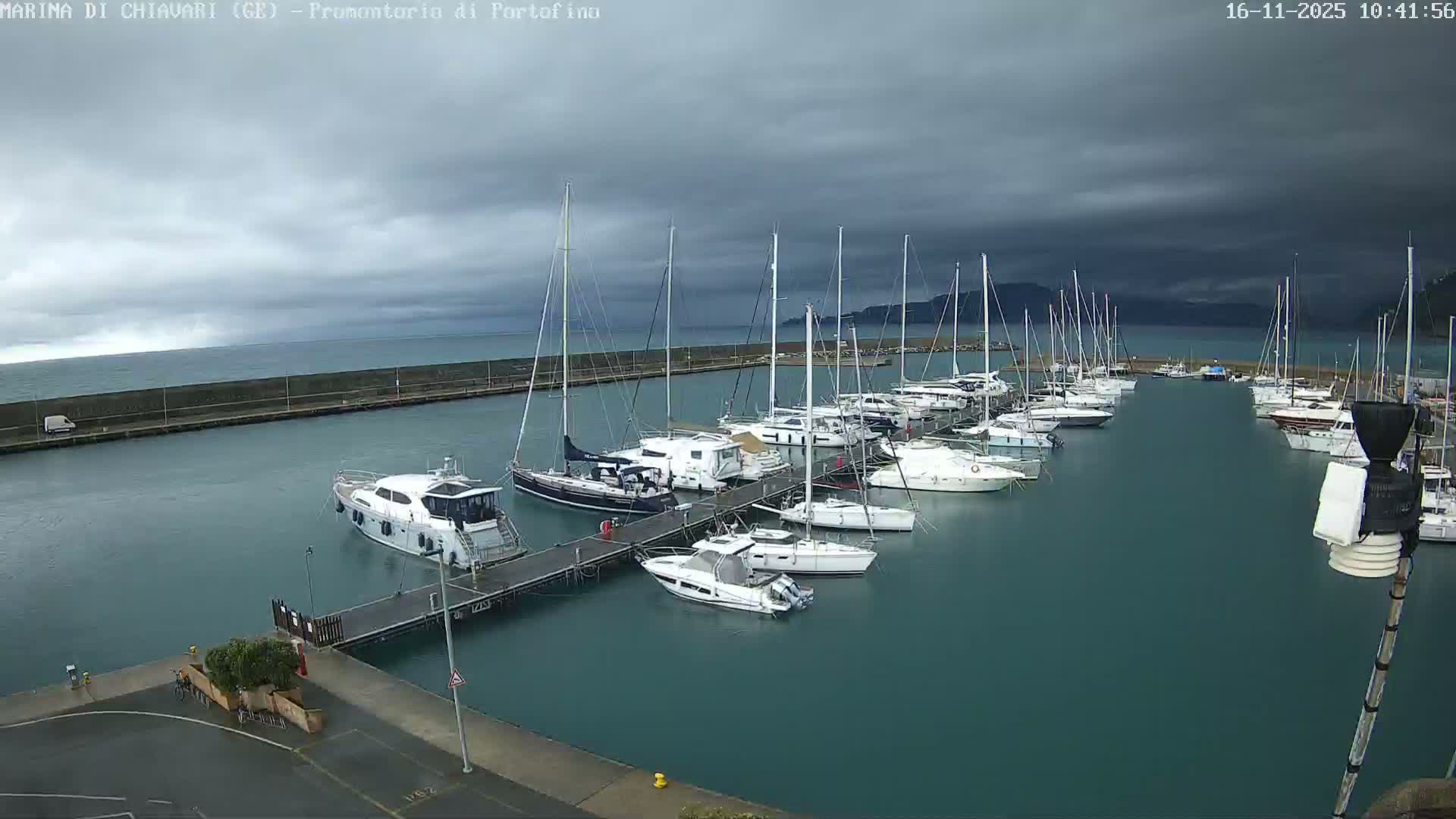 Numerous sailboats and yachts are moored in a teal-colored marina under a heavily overcast and stormy sky, with dark clouds looming over distant coastal hills.