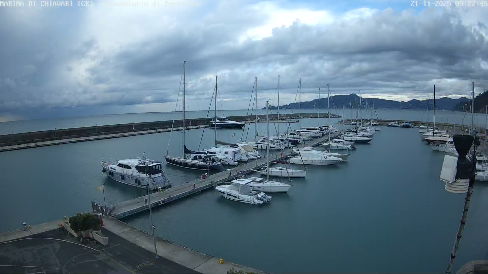 Numerous sailboats and motor yachts are docked in a marina under a heavily overcast sky, with a long breakwater extending into the sea and distant hills lining the horizon.