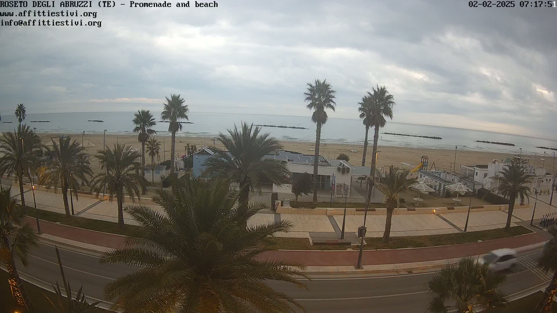 A mostly cloudy day overlooks a sandy beach with palm trees lining a paved promenade and road, a car driving by.
