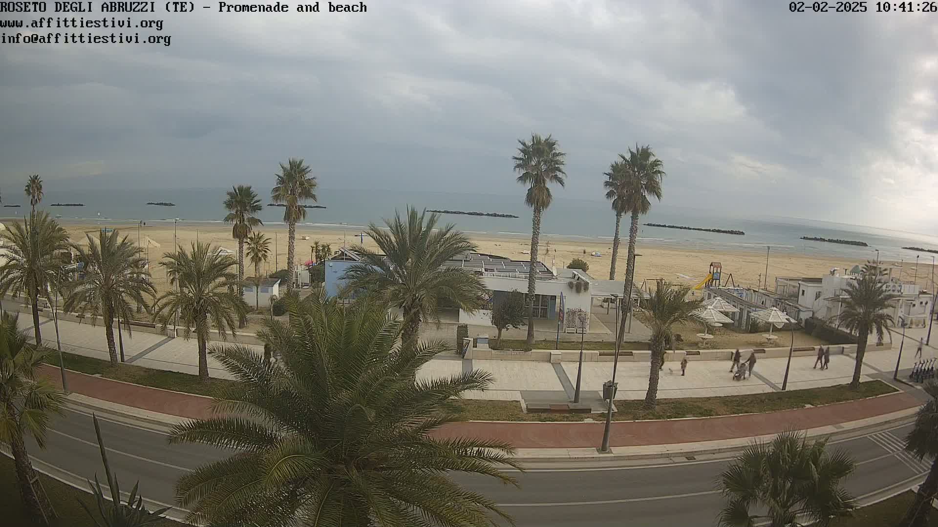 A seaside promenade lined with palm trees overlooks a sandy beach and calm ocean under a cloudy sky.