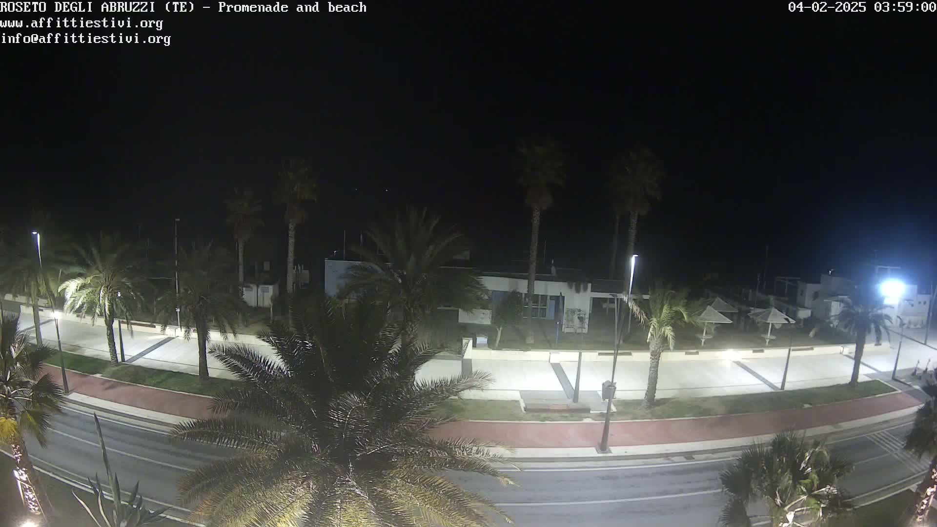 A nighttime, overhead view of a mostly empty promenade and beach, lined with palm trees and illuminated by streetlights.