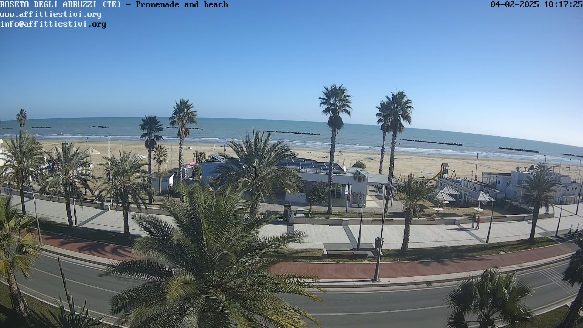 A beach with calm, blue water under a clear sky is viewed from above, showing palm trees lining a paved promenade next to beachside buildings and a sandy shore.
