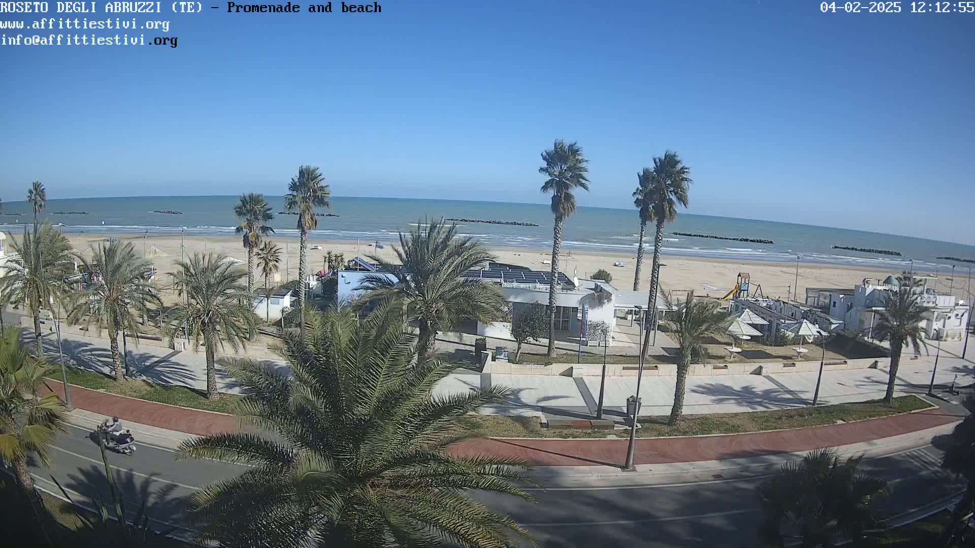 A sunny day at a beach promenade shows palm trees lining a road next to a sandy beach with calm ocean waves, some beach buildings, and a few people visible.