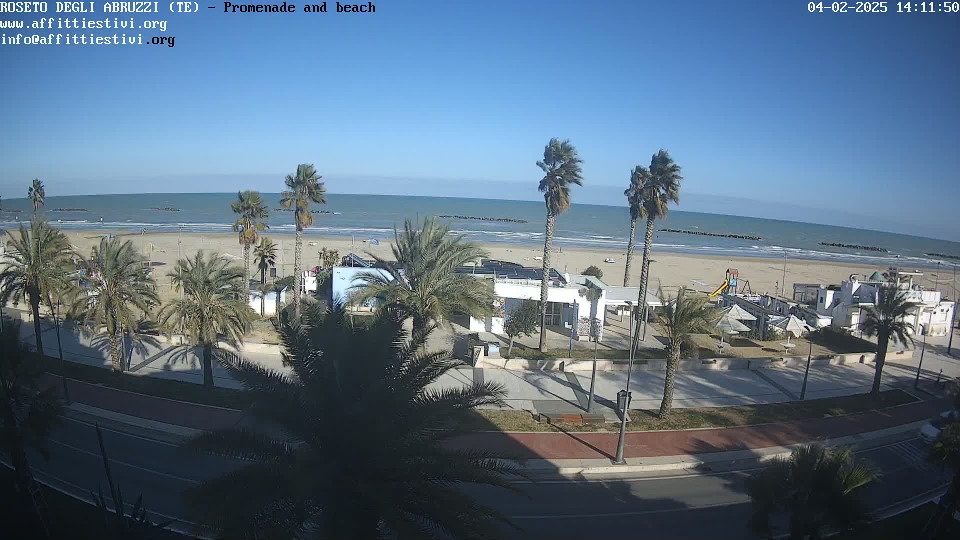 A beach with calm ocean waves under a clear blue sky, viewed from above a palm-tree lined promenade with beachside buildings.