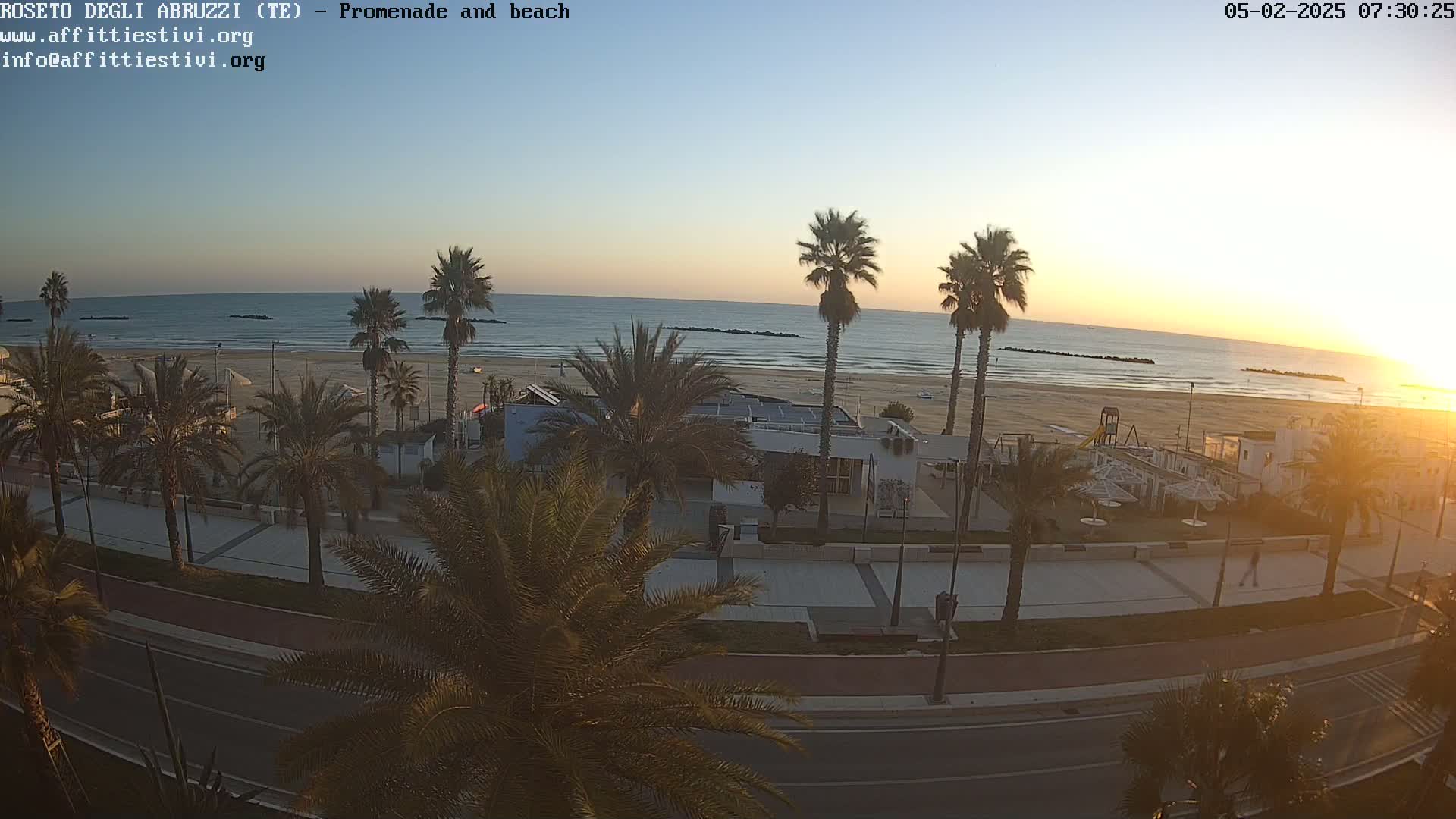 A sunrise illuminates a beach with palm trees lining a promenade and road next to a calm ocean.