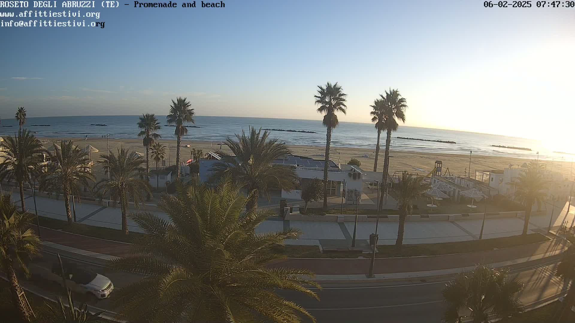 A beach with calm water under a clear, light-blue sky is viewed from above, showing a promenade lined with palm trees, buildings, and a roadway with a car.