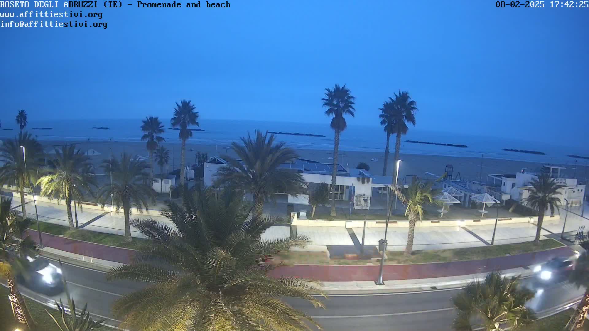 A beach at twilight, with palm trees lining a promenade beside a calm ocean.