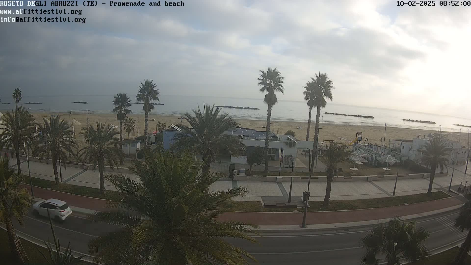 A mostly cloudy day overlooking a sandy beach with palm trees lining a promenade and a road, featuring a small building and playground.