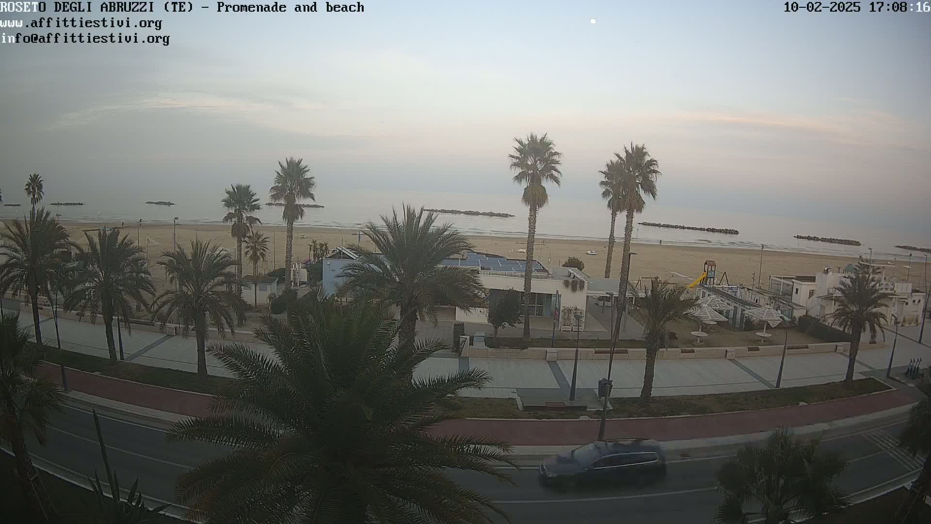 A beachfront promenade lined with palm trees, a sandy beach, calm ocean, and a car driving on the road beside it, under an overcast sky.
